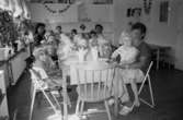 Midsommarfirande för barn och förskollärare som sitter inomhus i köket vid ett fint dukat bord. Barnen har kransar gjorda av papper i håret. Lunkentussen, Katrinebergs daghem 1992-93.