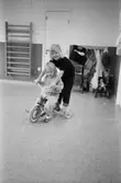 Två barn som sitter alternativt står på en trehjuling inomhus på Katrinebergs daghem. I bakgrunden ser man en ribbstege av trä uppsatt på väggen, några dockvagnar och filtar.