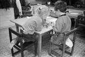 En flicka och en pojke, fotograferade bakifrån, sitter vid ett lågt bord och pysslar. I bakgrunden ser man leksaker och fler bord för barn. Katrinebergs daghem, 1992-93.