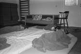 Tre barn ligger och sover middag på utlagda madresser på golvet. De har täcke på sig. I bakgrunden ser man sittkuddar, en ribbstol, soffa och stol. Katrinebergs daghem.