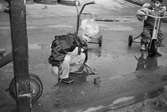 Robin leker i en vattenpöl med en spade i handen. Han är klädd i regnkläder och gummistövlar. Robin har använt en trehjuling som står bakom honom. I bakgrunden ser man ett barn som sitter på en annan trehjuling. Hoppetossan, Katrinebergs daghem, 1992.