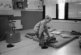 En pojke lutar sig ned och håller på en filt som ligger på en madrass på golvet. På madrassen ligger en kudde. Kanske dags för en liten sovstund? Katrinebergs daghem, 1992.