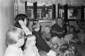 Två förskollärare och flertalet småbarn sitter tillsammans i en cirkel på golvet. Utanför rummets fönster kan man se en annan närliggande byggnad. Katrinebergs daghem, 1992.