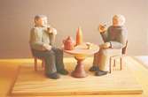 Två gubbar sitter på var sin stol vid ett runt bord och dricker ur varsitt ölkrus. På bordet står en kaffekittel, en sockerskål och en vinflaska. Harry Bergmans 