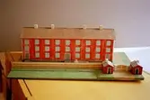 Ett rött bostadshus med tio fönster i rad, tre våningar högt, och tre entréer. På taket finns fyra skorstenar. Vid sidan av ligger två mindre stugor. Huset är en kopia av 