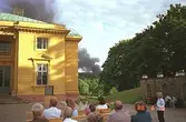 Publik som sitter på bänkar framför Gunnebo slott och lyssnar till en sångkör. Man kan se mörkgrå brandrök i fjärran, som troligtvis kommer från en grill.