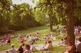 Människor som sitter i gräset och lyssnar på Lasse Berghagen (ej i bild) som sjunger på en tillfälligt uppbygd scen. Gunnebo slotts park, 29 maj 1988.