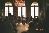 Människor som lyssnar till en konsert inomhus på Gunnebo slott. Personerna som spelar instrumenten är iklädda  historiserande dräkter. I bakgrunden ser man en flygel och i taket hänger en stor, pampig kristallkrona.