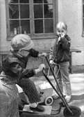 En pojke åker på en sparkcykel och en flicka står med en barnvagn på gårdsplanen utanför huset. Holtermanska daghemmet juni 1973.