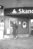 En kille står framför en Bankomat som är placerad utanför Skandinaviska Enskilda banken. Mölndalsbro i dag - ett skolpedagogiskt dokumentationsprojekt på Mölndals museum under oktober 1996. 1996_1061-1076 är gjorda av högstadieelever från Kvarnbyskolan 9C, grupp 3. Se även 1996_0913-0940, gruppbilder på klasserna 1996_1382-1405 samt bilder från den färdiga utställningen 1996_1358-1381.