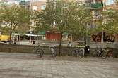 Cyklar uppställda vid Alberts torg. På torget ser man torghandel och en scen. Mölndalsbro i dag - ett skolpedagogiskt dokumentationsprojekt på Mölndals museum under oktober 1996. 1996_1301-1319 är gjorda av högstadieelever från Åbyskolan 8A, grupp 4. Se även 1996_0913-0940, gruppbilder på klasserna 1996_1382-1405 och bilder från den färdiga utställningen 1996_1358-1381.