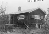 Fotografens namn står som regel inte utsatt, men fotograf är oftast Kalle själv, ibland någon tidningsfotograf och ibland någon klubbkamrat. Under bilden står: STUGAN. På albumets fotografi står: MCK-STUGAN. Den blev klar 1935 och står mitt inne iskogen, mot Ubbhult till. Allt material måste bäras dit, även järnspisen. Den bars dit av en klubbmedlem, som bar den på ryggen. Stugan finns också på MMF 1997:3510.