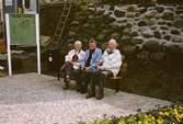 Tre personer sitter på en parksoffa vid Lyktplatsen, Kvarnbydagen 25/4 1993. Tredje person från vänster är Vigon Olsson. Gatlyktan (ej i bild) invigs. Till vänster ses en skylt över 