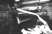 Två män vid en maskin för papperstillverkning, 1980-tal. Mannen som drar i papperet är Kent-Erik Pettersson.
Bilden ingår i serie från produktion och interiör på pappersindustrin Papyrus.