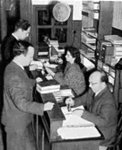 Stadsbibliotekarie Edvin Trettondal (glasögonprydd, sittandes till höger) i gamla biblioteket på Kvarnbygatan 49 (idag: Samuel Norbergsgatan 2), okänt årtal. Avfotograferad ur 