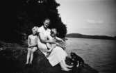 En kvinna med två barn och en liten hund sitter på en klippa, troligtvis vid Tulebosjön cirka 1930.