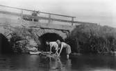 Sommar i Lindome i början av 1940-talet. Två kvinnor skurar mattor ståendes i vatten (troligtvis Lindomeån). I bakgrunden ses en stenbro.