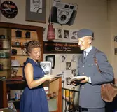 Brevbärare iklädd postens uniform från 1962 års
beklädnadsreglemente, överlämnar post till kvinnlig expedit i en
fotobutik. Brevbäraren på bilden är med stor sannolikhet Owe Jonsson.
Owe Jonsson var brevbärare i Växjö och vann EM-guld i Belgrad den 16
september 1962 i löpning 200 meter, bara 13 dagar senare omkom han i
en bilolycka.