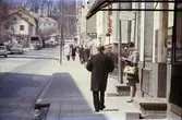 Gångtrafikanter och en kvinna som står och väntar på bussen vid Frölundagatan i Mölndal, 1960-tal.