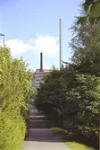 Asfalterad gångväg, grönska, takåsar och två skorstenar fotograferade nerifrån gångbanan vid Kvarnbygatan i Mölndals Kvarnby. Den vänstra röda skorsten sitter på 