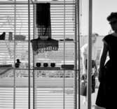 H55, Helsingborgsutställningen
Interiör, utställningshyllor mot glasvägg med persienn, del av kvinna i motljus