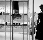 H55, Helsingborgsutställningen
Interiör, utställningshyllor mot glasvägg med persienn, del av kvinna i motljus