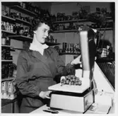 Bolstad, Lillebyn. Inger Eriksson står bakom disken och väger vindruvor på en våg.