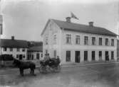 Häst och vagn framför lägenhetshus med affärer vid Vetlanda torg.