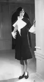 Modell i mörk kappa med krage och ärmslut i vit päls, hatt och pumps.