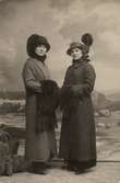 Två kvinnor stående framför målad fond. Kvinnorna iklädda ytterkläder, kappa, muff och hatt.