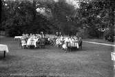 Män klädda i vita mössor och deras familjer har samlats, ute på en gräsmatta med träd i bakgrunden, kring dukade långbord.