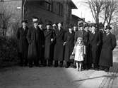 Män klädda i rock och hatt står på rad på en trottoar. Framför dem står en liten pälsklädd flicka med handväska.
