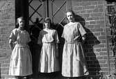 Tre unga kvinnor framför en dörr i ett tegelhus. Längst till höger står fotograf Gustav Anderssons syster Elsa.