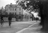 Stenhus kantar gatan där två män cyklar, vid andra sidan växer stora träd.