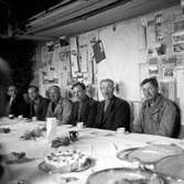 Sex män vid ett kaffebord. I bakgrunden en anslagstavla med tidningsartiklar om det nya flygfältet. Flygfältet började byggas år 1929 som stadskommunalt nödhjälpsarbete. Sex år senare skedde den högtidliga invigningen i Jönköping.