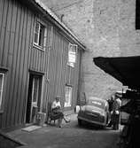 En bakgård vid Smålandsgatan i Jönköping. På stolen sitter Inga-Britt Friman, innehavarinna av Ajax hattaffär. Vid bilen står bilförsäljare Oscar Jonsson och Jönköpingsprofilen Häggman med ett paket under armen. Till höger syns delar av Elander & Lundbergs järnlager.