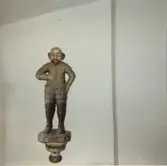 Skulptur. Knätte En mansfigur med mustasch och pipskägg. Klädd som en soldat, det liknar en rustning. Har hållit något i höger hand, hålet för fästet visar på det, men föremålet är borta. Kanske var det en fana?