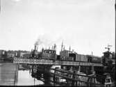 Ett tåg på UVHJ-banan passerar den gamla svängbron i anslutning till järnvägsstationen i Vänersborg.
Foto fr. ca 1915.