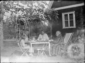 Fyra personer dricker kaffe i trädgården. Stugan sannolikt belägen vid Nordkroken, Vargön.