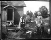 Olga, född Olsson, Forshälla 1891-05-12 och Beda Jonsson, född Björn, Bäve 1892-12-20 tvättar kläder framför boningshuset på Torpet (Arnstorp) i bakgrunden syns också en del utav källarbyggnaden. I vaggan sitter Sven Jonsson, Hjärtum 1915-05-14.
