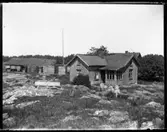 Vy över boningshuset på Arnstorp, det gamla boningshuset skymtar på i bakgrunden och den gamla ladugården syns till vänster på bilden. På bilden syns också flaggstången och timret till ny ladugård på Arnstorp som laftats upp innan den skall monteras på sin rätta plats.