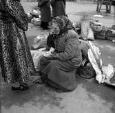 Denna bild är troligen från Stockholm dit fotograf Gustav Andersson ibland reste. På ett torg en sitter en äldre kvinna omgiven av väskor och påsar, en med texten 