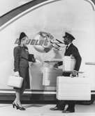 Kvinnlig resenär i dräkt, hatt med flor och handskar med resväska i handen, vid flygplanspersonal med resväskor. Douglas Aircraft.