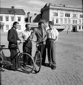 På Östra torget i Jönköping står fyra män med två cyklar. De två i mitten är från vänster Gösta Fröding och Olle Lindberg. I bakgrunden syns Amerikahuset.