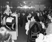 Kvinna demonstrerar skönhetsprodukter från Dorothy Gray för publik på Nordiska Kompaniet.