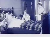 Varbergs gamla sjukhus, kv Lasarettet. Två sjuksköterskor vid Mathilda Ranchs sjuksäng 1908. Mathilda håller i ett glas och vid sidan av sängen står en stor blomsterbukett med hyacinter och tulpaner.