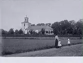 En kvinna och två flickor promenerar. Slöinge kyrka och kyrkogård i bakgrunden. Bild utan år.