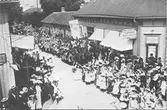 Barnens Dag firande i Varberg 1906. Vy över ett parti av Drottninggatan. Trähusbebyggelse. Åskådare, barn och vuxna, kantar gatan för att beskåda karnevalståget.