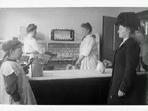 Interiör från en mjölkaffär år 1912 på Prästgatan, kv Guldsmeden, Varberg. Innanför disken står expediterna Josefina Johansson och Alma Karlsson. Flickan i keps heter Herdis Sandgren men namnet på kunden till höger är okänt.