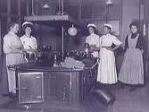 Kökspersonal i köket på länslasarettet i Varberg ca 1913. Kvinnorna står med kastruller runt en i rummet fritt stående spis. Vedträn ligger ovanpå ena hörnet.